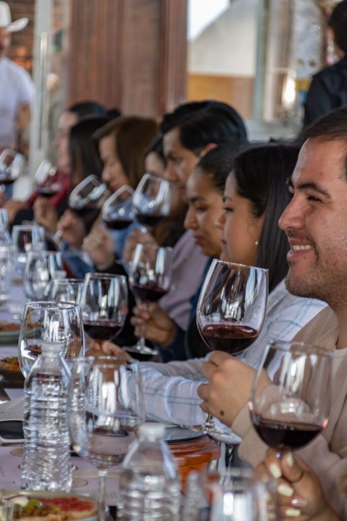 A diverse group participates in a wine tasting, enjoying red wine at a communal table indoors.